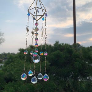 May include: A hanging suncatcher with a geometric gold frame and iridescent crystal beads. The suncatcher features a diamond-shaped top with dangling chains, sun-shaped charms, and multiple crystal spheres. The crystals reflect a rainbow of colors.