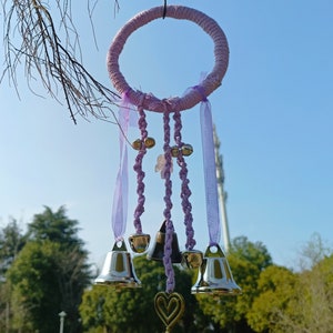 Puede incluir: Un atrapasueños de macramé morado y blanco con campanas colgando. El atrapasueños está hecho de hilo y tiene un anillo de madera en la parte superior.