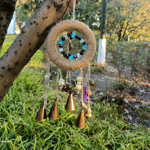 Puede incluir: Un atrapasueños marrón con cuentas azules y negras colgando de una rama de árbol. El atrapasueños tiene una variedad de campanas y encantos colgando de él, incluyendo pequeñas campanas de metal, cuentas de madera y un cristal morado.