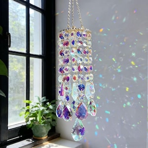 Könnte beinhalten: Ein hängender Kristall-Sonnenfänger mit einer goldfarbenen Kette. Der Sonnenfänger besteht aus schillernden Kristallen in Lila, Blau und Klar. Die Kristalle sind in einem kaskadenartigen Muster angeordnet, mit größeren tropfenförmigen Kristallen unten.