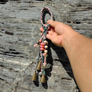 Może przedstawiać: Pleciony sznurek w kolorze różowym i zielonym z pętlą na górze. Sznurek ma przymocowane małe dzwoneczki i koraliki. Koraliki są różowe, białe i czarne.