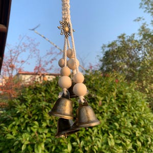 Peut inclure: Un carillon éolien suspendu, composé de trois cloches en bronze, de perles en bois et d'un charm en forme de soleil. Le carillon est suspendu à une corde en macramé. Le fond est composé de verdure et d'un ciel bleu clair.