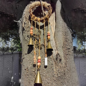 May include: A brown twig wreath with a beaded and bell wind chime hanging from it. The chime features small bells, beads, and glass vials. The wreath is hanging on a tree trunk.