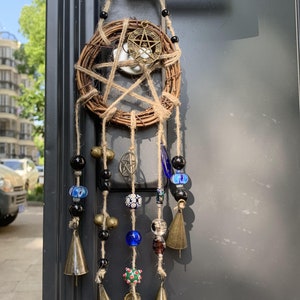 Puede incluir: Un atrapasueños tejido de color marrón con un amuleto de pentagrama y campanas colgando. El atrapasueños está decorado con cuentas de vidrio negras, azules y transparentes.