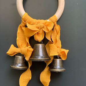 Puede incluir: Un anillo de madera con tres campanas de latón colgando de él. Las campanas están unidas a cintas de tela amarilla.