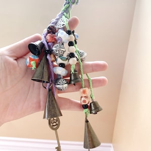 Puede incluir: Un cordón trenzado morado, verde y blanco con una variedad de encantos, incluyendo campanas, cuentas y una llave. Los encantos están enhebrados en el cordón y cuelgan de él.