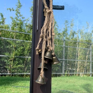 May include: A decorative wind chime featuring small, antique brass bells suspended from a natural jute rope. The bells are of varying sizes and hang in a cluster. The chime is attached to a dark brown post, with a green lawn and blue sky in the background.