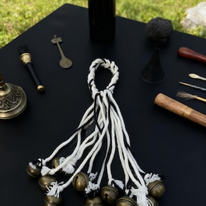 Könnte beinhalten: Eine Ansammlung kleiner Messingglocken, die an weiß-schwarzen geflochtenen Schnüren befestigt sind. Die Glocken sind zusammengefasst, wobei die Schnüre von einer zentralen Schlaufe ausgehen. Die Anordnung befindet sich auf einer schwarzen Oberfläche, mit anderen dekorativen Gegenständen im Hintergrund.