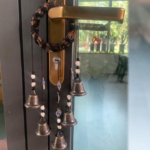 Puede incluir: Un carillón negro y blanco con una corona tejida en la parte superior y cinco pequeñas campanas colgando de él. Las campanas son plateadas con dijes de ojo turco azul y blanco. El carillón está colgado en una puerta.