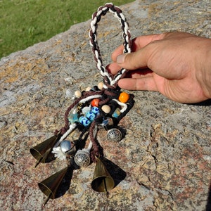 以下が含まれることがあります： 編組された紐のループ、カラフルなビーズ、4つの小さなベルが付いた手作りチャーム。チャームは茶色、白、黒の紐で作られており、青、オレンジ、白のビーズが特徴です。