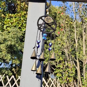 May include: A black dreamcatcher with a pentagram charm and blue evil eye beads hanging from it. The dreamcatcher is adorned with small, gold-colored bells.