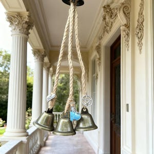 May include: A hanging wind chime featuring three bronze-colored bells suspended from a rope. Each bell has a decorative charm, including a heart, a potion bottle, and a pentagram. The bells are hanging on a porch.