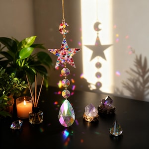 Puede incluir: Un atrapasoles de temática celestial con un diseño de estrella y media luna, adornado con cristales de colores. El adorno en forma de estrella está adornado con una variedad de gemas de colores. Un cristal en forma de lágrima cuelga debajo.