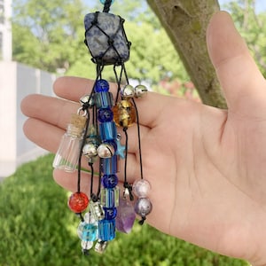 Peut inclure: Un charme en cristal bleu et noir avec un petit flacon en verre, des perles bleues et dorées et des clochettes argentées qui pendent.