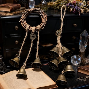 May include: A rustic, witchy aesthetic featuring a woven wreath and a set of hanging bells. The bells are bronze-colored and suspended from twine, with a pentagram charm. An open book and crystals are also visible.