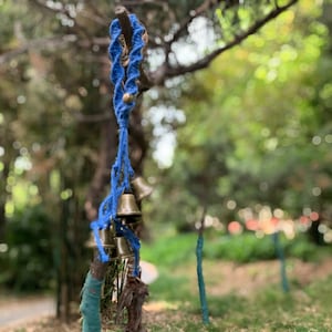 May include: A blue rope with a series of small, gold-colored bells hanging from it. The bells are tied to the rope with knots. The rope is hanging from a tree branch.
