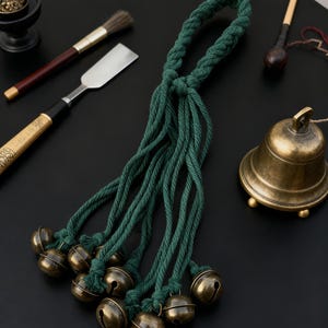 Könnte beinhalten: Ein grünes Seil mit mehreren Strängen, die jeweils in einer kleinen, antiken Messingglocke enden. Die Glocken sind zusammengefasst und das Seil ist oben verknotet. Weitere Artikel sind eine kleine Messingglocke, ein Pinsel und ein Metallwerkzeug.