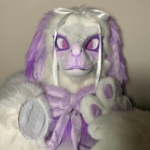 May include: A creature mask featuring white and purple faux fur, a textured white reptilian face, and purple eyes. The mask is adorned with a white satin ribbon bow on top and a large purple fabric bow at the neck, accompanied by matching white and purple paw gloves.
