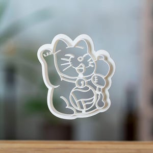 Może przedstawiać: Biała plastikowa foremka do ciastek w kształcie japońskiego kota Maneki Neko, z podniesioną łapą i trzymającego monetę. Kot ma uśmiechniętą twarz i nosi obrożę z dzwoneczkiem. Foremka leży na drewnianej powierzchni.