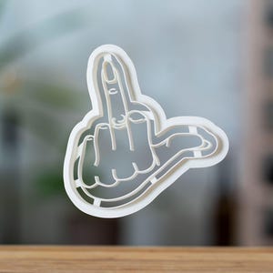 May include: A white plastic cookie cutter in the shape of a hand giving the middle finger. The cutter is a single piece with a detailed outline of the hand and finger.