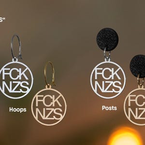 Puede incluir: Dos pares de pendientes con colgantes circulares con el texto FCK NZS. El par izquierdo consiste en aros plateados y dorados. El par derecho presenta pendientes de bot&oacute;n negros texturizados unidos a colgantes circulares plateados y dorados.