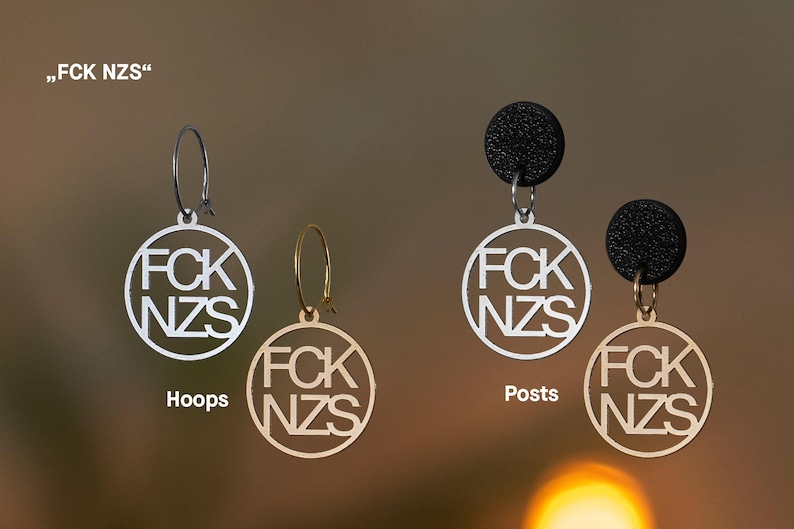 Puede incluir: Dos pares de pendientes con colgantes circulares con el texto FCK NZS. El par izquierdo consiste en aros plateados y dorados. El par derecho presenta pendientes de bot&oacute;n negros texturizados unidos a colgantes circulares plateados y dorados.
