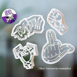 Puede incluir: Cuatro cortadores de galletas de plástico blanco de varias formas. Uno deletrea "RIOT!!!", otro es un puño con un símbolo femenino, y los otros son un puño y una mano haciendo un gesto obsceno. Un logotipo morado y blanco está en la esquina superior izquierda.