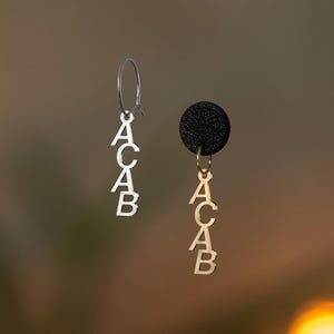 Könnte beinhalten: Ein Paar silberne und goldene Ohrringe mit den Buchstaben "ACAB", die daran hängen. Die Ohrringe befinden sich auf einem braunen Hintergrund.