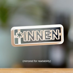 Puede incluir: Un letrero rectangular blanco con la palabra alemana "INNEN" en letras negritas sin serifa. El letrero incluye un símbolo médico y está reflejado para facilitar la lectura. El fondo está borroso.