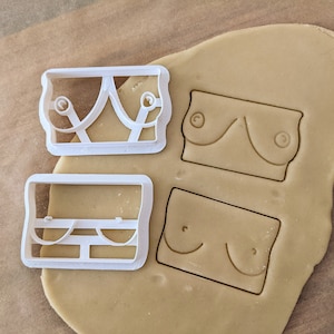 Puede incluir: Dos cortadores de galletas de plástico blanco sobre una superficie marrón claro. Los cortadores tienen forma de sujetador. Un cortador tiene un diseño simple, mientras que el otro tiene un diseño más intrincado con una correa decorativa.