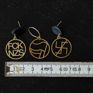 Puede incluir: Tres colgantes circulares de metal dorado con enganches negros, colocados sobre una cinta m&eacute;trica blanca que muestra una anchura de unos 10 cm. Los colgantes presentan dise&ntilde;os recortados que incluyen el texto FCK NZS y s&iacute;mbolos.