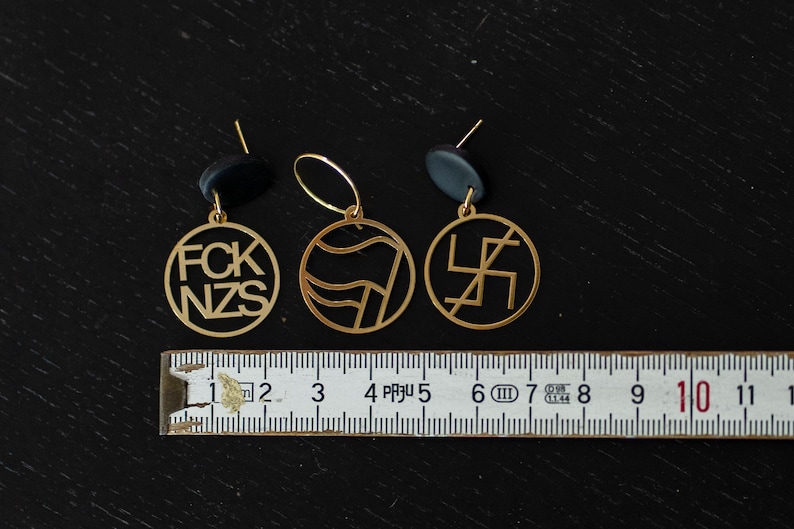 Puede incluir: Tres colgantes circulares de metal dorado con enganches negros, colocados sobre una cinta m&eacute;trica blanca que muestra una anchura de unos 10 cm. Los colgantes presentan dise&ntilde;os recortados que incluyen el texto FCK NZS y s&iacute;mbolos.
