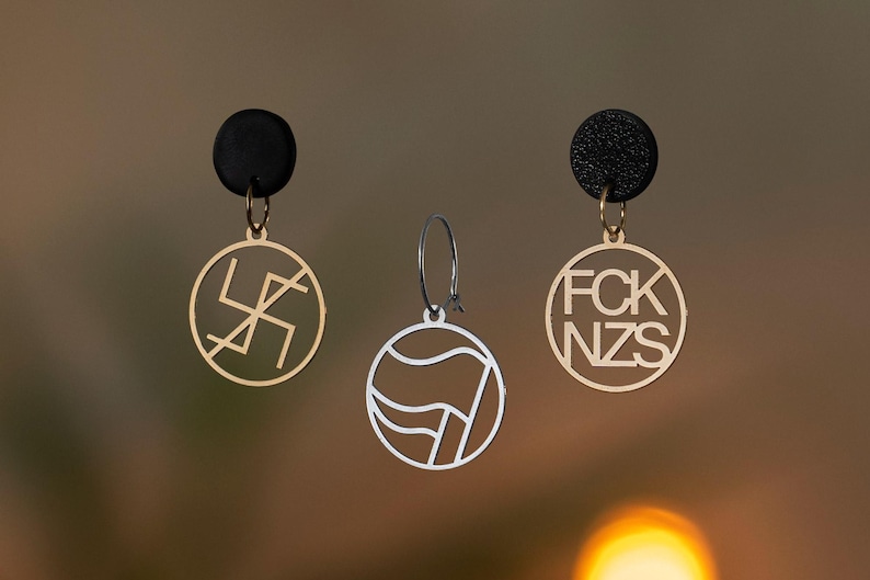 Puede incluir: Tres pendientes: dos con postes circulares negros y colgantes circulares dorados, uno con un s&iacute;mbolo tachado y el otro con el texto FCK NZS. El tercero es un colgante circular plateado en un aro con un dise&ntilde;o de bandera.