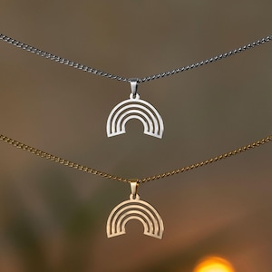 Może przedstawiać: Dwa naszyjniki z wisiorkami w kształcie tęczy. Górny naszyjnik ma srebrny wisiorek, a dolny złoty.