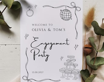 May include: A white engagement party sign with black line art illustrations of a diamond ring, disco ball, champagne glasses, and bows. The text reads "Welcome to Olivia & Tom's Engagement Party 15.08.2027". Decorative sprigs of greenery and dried flowers are in the background.