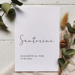 May include: A white card with black text that reads "Santorini" in cursive, "Elizabeth & Tom" and "15-08-2026".
