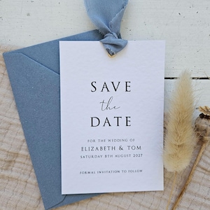 May include: A wedding save-the-date card with a blue envelope and ribbon. The white card reads "SAVE the DATE" and includes the names Elizabeth & Tom and the date 8th August 2027. A formal invitation will follow.