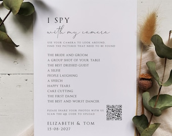 May include: A white wedding activity card with the text "I Spy with my camera" and a list of photo prompts. The card includes a QR code and the names Elizabeth & Tom with the date 15-08-2027. The card is decorated with greenery.