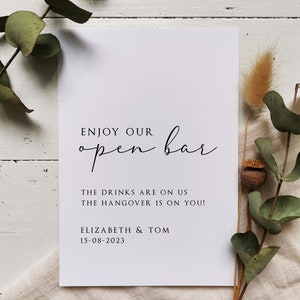 May include: White card with black text that reads "Enjoy our open bar. The drinks are on us. The hangover is on you! Elizabeth & Tom 15-08-2023". The card is on a white linen surface with eucalyptus branches and dried pampas grass.