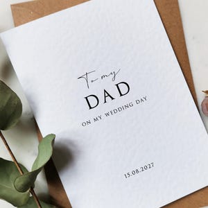 May include: A white card with the text "To my DAD ON MY WEDDING DAY" in black script. The date "15.08.2027" is printed at the bottom. The card is on a brown envelope, with green leaves and dried flowers in the background.