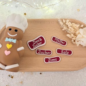May include: A gingerbread man chef figurine with a white hat and bow tie, next to wooden labels with German text. The labels are red with white borders and text. A wooden serving tray holds the items.