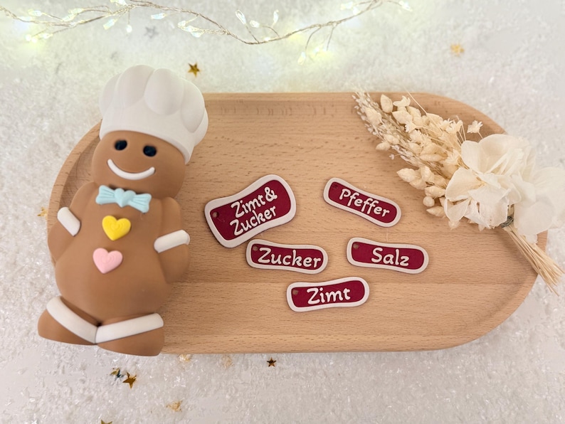 May include: A gingerbread man chef figurine with a white hat and bow tie, next to wooden labels with German text. The labels are red with white borders and text. A wooden serving tray holds the items.