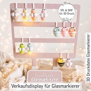 Puede incluir: Un soporte de exhibición rosa claro con tres barras horizontales que sostienen varias figuras pequeñas y coloridas con forma de abeja. El soporte tiene una placa base con el texto "Leuchtende Glasmarkierer" y un letrero "STL & 3MF für 3D Druck".