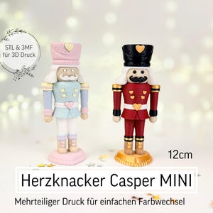 Puede incluir: Dos figuras de cascanueces en miniatura, una en azul pastel y rosa, la otra en rojo y negro, de pie una al lado de la otra. Cada una tiene un detalle en forma de corazón en el sombrero. El texto "Herzknacker Casper MINI" se muestra debajo, con una altura de 12 cm.