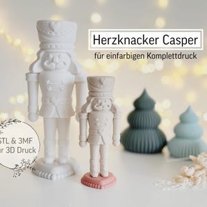 May include: White and light pink nutcracker figurines and decorative Christmas trees. The image includes the text "Herzkknacker Casper" and "STL & 3MF für 3D Druck". The nutcrackers are standing on a white surface.