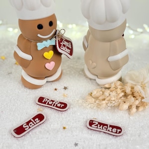 May include: Two gingerbread chef figurines with white chef hats, bow ties, and heart accents. The figurines stand on a white surface with small red and white labels that read "Salz", "Pfeffer", and "Zucker".