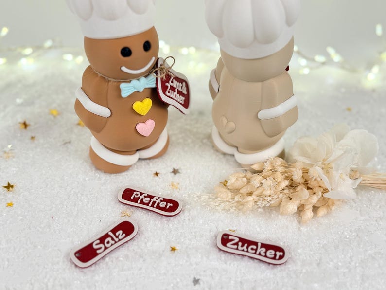 May include: Two gingerbread chef figurines with white chef hats, bow ties, and heart accents. The figurines stand on a white surface with small red and white labels that read "Salz", "Pfeffer", and "Zucker".