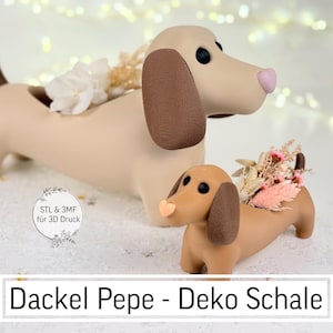 May include: Tan dachshund-shaped decorative bowls, one large and one small, with brown ears and tails, and pink noses. The bowls are filled with dried flowers. The text "Dackel Pepe - Deko Schale" is at the bottom.
