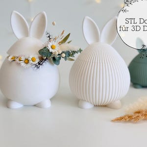 May include: White ceramic bunny-shaped containers with upright ears. One bunny has a floral crown. The other has vertical lines. A green bunny-shaped container is in the background. Text in German says "STL File for 3D Print."