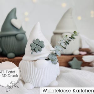 May include: Three gnomes, one white with a four-leaf clover on its hat, one green with a heart on its hat, and one gray with a sprig of eucalyptus. The gnomes are sitting on a wooden surface with the text "STL Datei für 3D Druck" and "Wichteldose Karlchen" below them.